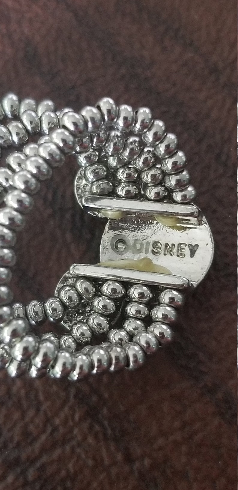May include: Close-up of a silver-toned bracelet with a beaded design. The bracelet has a clasp with the word "DISNEY" stamped on it. The beads are small and round, and the bracelet appears to be vintage.