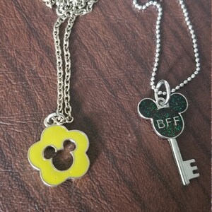 May include: Two silver-toned necklaces with pendants. One pendant is a yellow flower shape with a Mickey Mouse silhouette. The other is a key shape with a Mickey Mouse head and the letters "BFF" in green glitter.