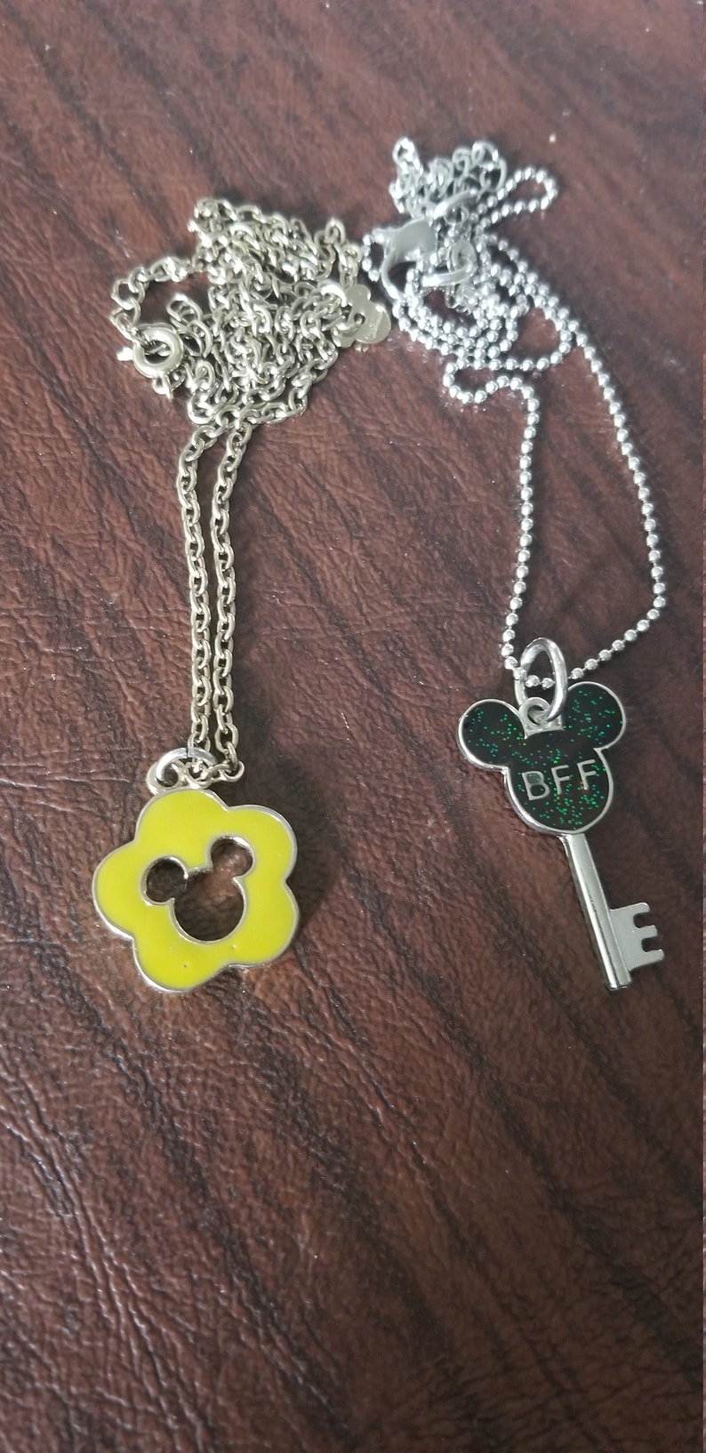 May include: Two silver-toned necklaces with pendants. One pendant is a yellow flower shape with a Mickey Mouse silhouette. The other is a key shape with a Mickey Mouse head and the letters "BFF" in green glitter.