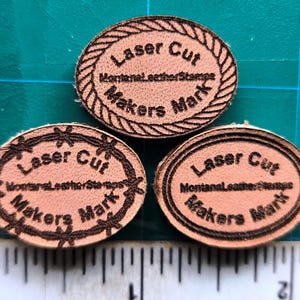 May include: Three oval-shaped leather stamps with a brown background and black text. The text on the stamps reads "Laser Cut", "Montana Leather Stamps", and "Makers Mark".