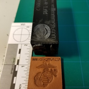 May include: A black rubber stamp with the text "www.vollkommen.com" and the words "Sga-R-1.rld 90909.2" on the side. The stamp is next to a brown leather patch with the text "www.vollkommen.com" and a stylized image of the US Marine Corps emblem.