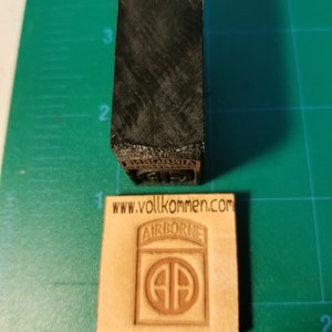 May include: A brown leather stamp with the text "AIRBORNE" and the 82nd Airborne Division insignia, a circle with the letters "AA" inside. The stamp is on a green gridded surface. The text "www.vollkommen.com" is printed below the stamp.