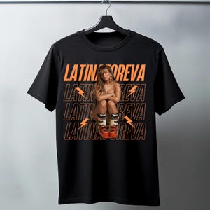 May include: Black t-shirt with the text "LATIN REVA" in orange, repeated four times. A woman is centered on the shirt, wearing ski boots. The shirt is hanging on a black hanger.
