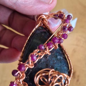 May include: A teardrop-shaped dark blue and green stone pendant wrapped in copper wire. The wire design features intricate coils and is accented with small, round, faceted magenta-colored beads along the side and top of the stone.