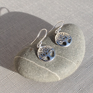 May include: A pair of silver earrings featuring a tree of life design. The earrings are hanging from a smooth grey stone.