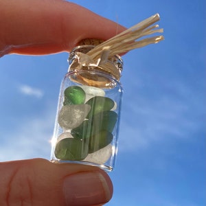 May include: A miniature glass bottle filled with green and white sea glass, held against a blue sky. The bottle has a cork stopper tied with raffia. The sea glass pieces are smooth and rounded, creating a natural aesthetic.