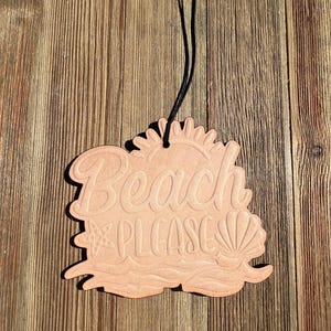May include: A light brown, beach-themed air freshener with the words "Beach PLEASE" in a decorative font. The design includes a sun, starfish, waves, and seashells. A black string is attached for hanging.