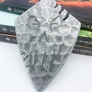 Puede incluir: Un colgante de forma triangular de color plateado con un diseño de calavera. La calavera está hecha de un material texturizado con forma de panal. El colgante es un artículo decorativo.