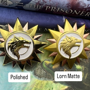 Puede incluir: Dos pines de metal de color dorado con un diseño de cabeza de lobo blanco en el centro. El pin de la izquierda está pulido y el pin de la derecha tiene un acabado mate. Los pines están etiquetados como "Polished" y "Lorn Matte".