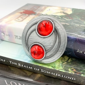 Puede incluir: Un medallón de metal plateado con dos piedras preciosas rojas engastadas en el centro. El medallón tiene un diseño circular con círculos concéntricos alrededor de las piedras.