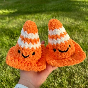 May include: Two crocheted orange and white traffic cone-shaped plush toys with smiling faces. The toys are held in a person's hand.