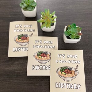 May include: Three greeting cards with the text "IT'S YOUR PHO-KING BIRTHDAY" and an illustration of a bowl of pho. The cards are beige with a cartoon image of pho. Three small white square pots with green succulents are also in the image.