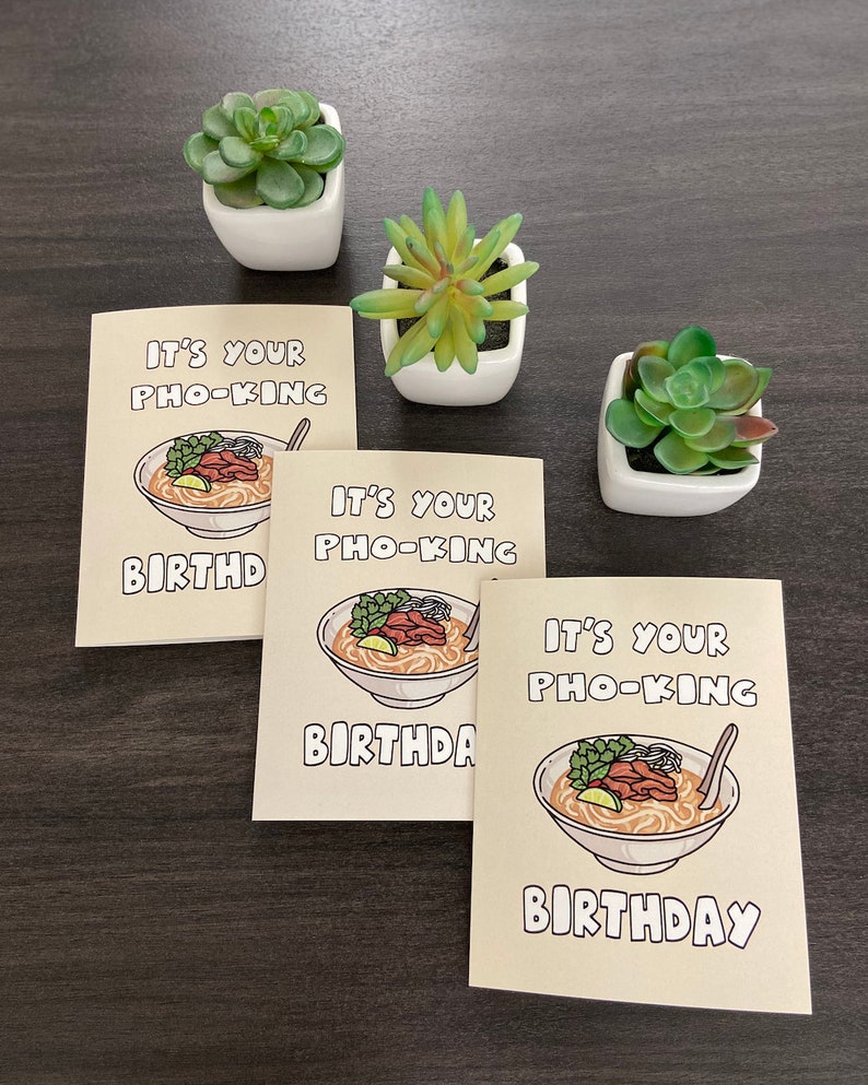 May include: Three greeting cards with the text "IT'S YOUR PHO-KING BIRTHDAY" and an illustration of a bowl of pho. The cards are beige with a cartoon image of pho. Three small white square pots with green succulents are also in the image.