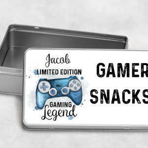 May include: A white metal tin with a blue watercolour gaming controller design. The text "Jacob Limited Edition Gaming Legend" and "Gamer Snacks" is printed on the tin.
