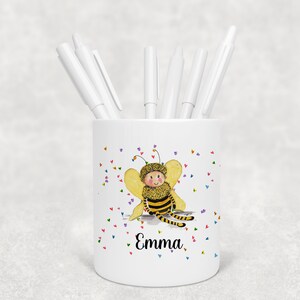 May include: White ceramic pencil holder featuring a cartoon bee design and the name "Emma" printed on the front. The bee is yellow and black with a smiling face and colourful hearts are scattered around it.