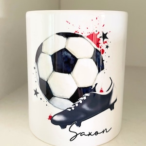 May include: White ceramic mug featuring a black and white football and a black football boot. Red and black paint splatters are in the background. The name "Saxon" is written in black cursive at the bottom of the mug.