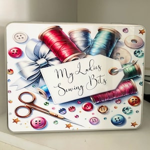 May include: A white tin box with a colourful design featuring spools of thread, buttons, and a tag that reads "My Ladies Sewing Bits".