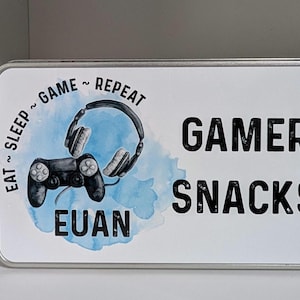 May include: Rectangular metal tin with a white background featuring a watercolour design of a black controller and headphones. Text reads "Eat ~ Sleep ~ Game ~ Repeat" and "Gamer Snacks" with the name "Euan".