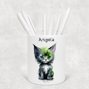 May include: White ceramic pencil holder featuring a watercolour illustration of a green and black cat. The holder is personalised with the name "Angela".