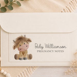 May include: Cream-coloured pregnancy notes folder with a flap closure. The front features a watercolour illustration of a Highland cow and the text "Baby Williamson PREGNANCY NOTES". A wooden baby toy and greenery are in the background.