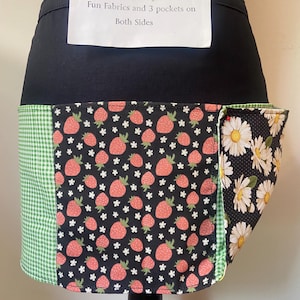 May include: Reversible apron with black waistband and three pockets. The apron features fun fabrics, including a green gingham, a black background with strawberries and white flowers, and a black background with white daisies. Text on the apron reads: "Reversible 2 Aprons in 1 Fun Fabrics and 3 pockets on Both Sides."