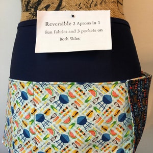 May include: Reversible apron with a navy blue waistband and a fun barbecue-themed print. The print features images of grills, watermelon, and skewers. The apron has three pockets and is reversible.