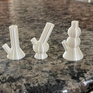 Może przedstawiać: Trzy białe ceramiczne bongi o różnych kształtach i rozmiarach. Bongi znajdują się na granitowym blacie.