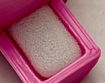 Könnte beinhalten: Eine leuchtend pinke Ringbox mit einer texturierten, 3D-gedruckten Außenfläche. Das Innere der Box verfügt über einen rechteckigen weißen Schaumstoffeinsatz, der dazu dient, einen Ring sicher zu halten. Der Deckel ist teilweise geöffnet.