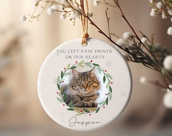 May include: White ceramic Christmas ornament with a photo of a tabby cat in a green wreath with red berries. The ornament has the text "You left paw prints on our hearts" and the name "Jasper" written below the photo.