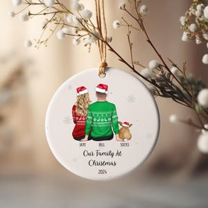 Peut inclure: Ornement de Noël rond en céramique blanche avec une illustration familiale. L'ornement représente un couple et un chien, tous portant des bonnets de Père Noël et des pulls de Noël. Le texte sur l'ornement se lit "Our Family At Christmas 2024".