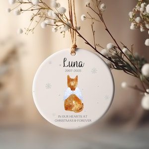 Könnte beinhalten: Runder, weißer Keramik-Weihnachtsschmuck mit einer Aquarellillustration einer rot getigerten Katze mit Engelsflügeln. Der Schmuck trägt den Namen "Luna" und die Daten "2007-2025" sowie den Text "IN OUR HEARTS AT CHRISTMAS & FOREVER".