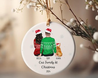 May include: A round white Christmas ornament with a family illustration. The ornament shows a couple and a cat, all wearing Santa hats and festive sweaters. The text "Our Family At Christmas 2024" is printed below the illustration, with names above.