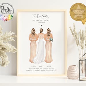 May include: A framed print with the text "To Our Sister On Your Wedding Day 08.06.2023." The illustration depicts three women in wedding attire. The print is in a light wood frame and features a gold seal that says "As Featured by Weddings."