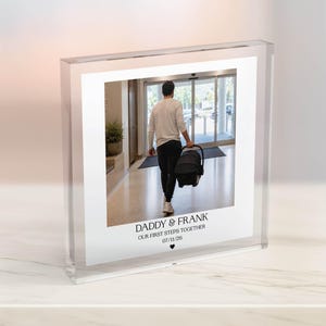 Puede incluir: Un bloque acr&iacute;lico transparente con una fotograf&iacute;a de una persona caminando con un portabeb&eacute;s. La foto tiene el texto "DADDY & FRANK OUR FIRST STEPS TOGETHER 07/11/26" en la parte inferior con un s&iacute;mbolo de coraz&oacute;n.