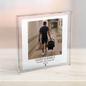 May include: A clear acrylic photo block with a photograph of a person walking down a corridor, carrying a baby carrier. The photo has text that reads "DADDY & JESSICA OUR FIRST STEPS TOGETHER 04.03.28".