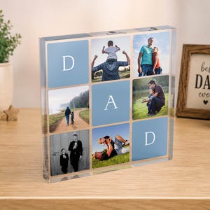 May include: A clear acrylic photo block with the word "DAD" in white letters. The block contains family photos and is displayed on a wooden surface. A framed picture with the text "Best Dad Ever" is visible in the background.
