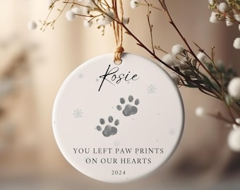 Könnte beinhalten: Runder weißer Keramikschmuck mit grauen Pfotenabdrücken und dem Namen "Rosie". Der Text lautet "YOU LEFT PAW PRINTS ON OUR HEARTS" und "2024". Aufgehängt an einer natürlichen Schnur, ideal als Haustiergedenken.