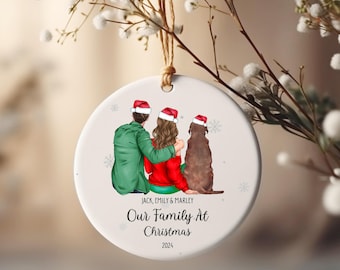 Può includere: Un ornamento natalizio rotondo con un'illustrazione familiare. Il design presenta una coppia e un cane, tutti con cappelli di Babbo Natale. L'ornamento ha il testo "Our Family At Christmas 2024" e i nomi "Jack, Emily & Marley".