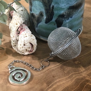 Puede incluir: Un infusor de té de plata con un mango en espiral y una bola de malla redonda. El infusor está sobre una superficie de madera con un trozo de coral y una taza de cerámica azul en el fondo.