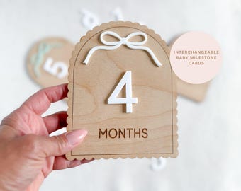 May include: Wooden baby milestone card for 4 months, with a white bow and scalloped edges. The number 4 and the word "MONTHS" are in white and brown. A pink circle with text "INTERCHANGEABLE BABY MILESTONE CARDS" is in the background.