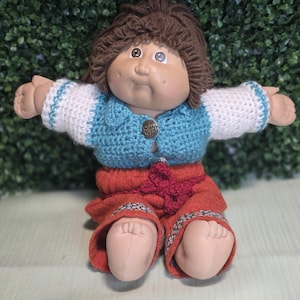Peut inclure: Une poupée avec des cheveux en laine brune et un visage rond aux joues marquées. Elle porte un gilet bleu au crochet sur une chemise blanche à manches longues avec des bordures bleues, et un pantalon rouille avec un lien décoratif à la taille.