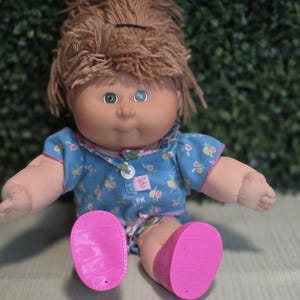 May include: A doll with light brown yarn hair, one green eye, and one blue eye. It wears a blue short-sleeved shirt with a small floral pattern, a white button, and a pink CPK tag. The doll has bright pink plastic shoes with textured soles.
