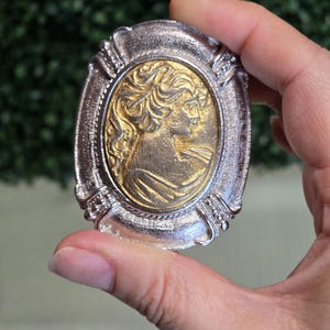 Peut inclure: Une broche de forme ovale présentant un relief doré d'un portrait de profil inséré dans un cadre argenté texturé. La figure centrale représente une personne aux cheveux ondulés vue de côté, entourée d'une bordure métallique en relief.