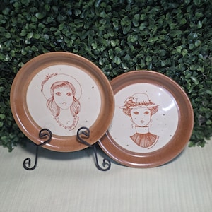 Peut inclure: Deux assiettes rondes en céramique avec un centre crème moucheté et un large bord marron. Chaque assiette présente une illustration au trait marron d'une personne portant un chapeau. L'assiette de gauche montre une personne aux cheveux longs.