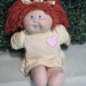 Peut inclure: Une poupée aux cheveux en laine rouge vif coiffés en deux couettes attachées par des rubans crème. La poupée a des yeux bleus, un visage rond et porte une robe jaune pâle à manches courtes avec un cœur rose appliqué et un ruban blanc.