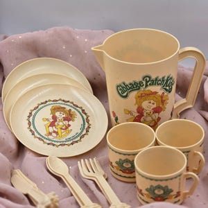 May include: A cream-coloured plastic Cabbage Patch Kids children's tea set including a pitcher, three plates, three mugs, two spoons, two forks, and two knives. Each piece features a graphic of a doll and floral accents with the text Cabbage Patch Kids.