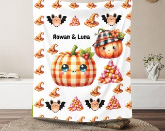 Könnte beinhalten: Eine weiße Halloween-Decke mit Cartoon-Kürbissen, Fledermäusen, Hexenhüten und Zuckermais. Die Kürbisse haben Karomuster und niedliche Gesichter. Der Text "Rowan & Luna" ist auf die Decke gedruckt.