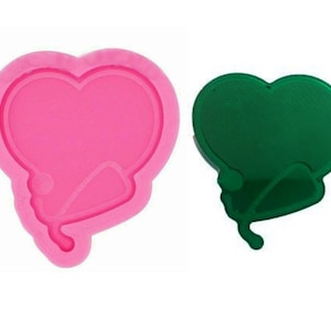 Puede incluir: Un molde de silicona rosa en forma de corazón con un estetoscopio unido a la parte inferior. Junto a él hay una fundición de resina verde del mismo diseño.
