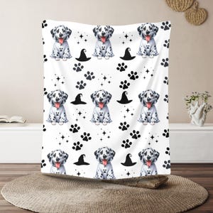 Könnte beinhalten: Weiße Decke mit einem sich wiederholenden Muster von Dalmatiner-Welpen in weißen Hemden mit schwarzen Punkten, schwarzen Hexenhüten, Pfotenabdrücken und Sternen. Die Welpen strecken die Zunge heraus. Die Decke wird auf einer weißen Oberfläche präsentiert.