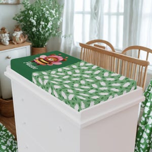 May include: A green changing pad cover with a white dresser. One side features a bee wearing a pink bonnet and the name "Phoebe". The other side has a pattern of white feathers and green leaves.